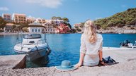 Junge Dame sitzt am Wasser vor einem Motorboot und blickt auf das bunte Fischerdorf Assos